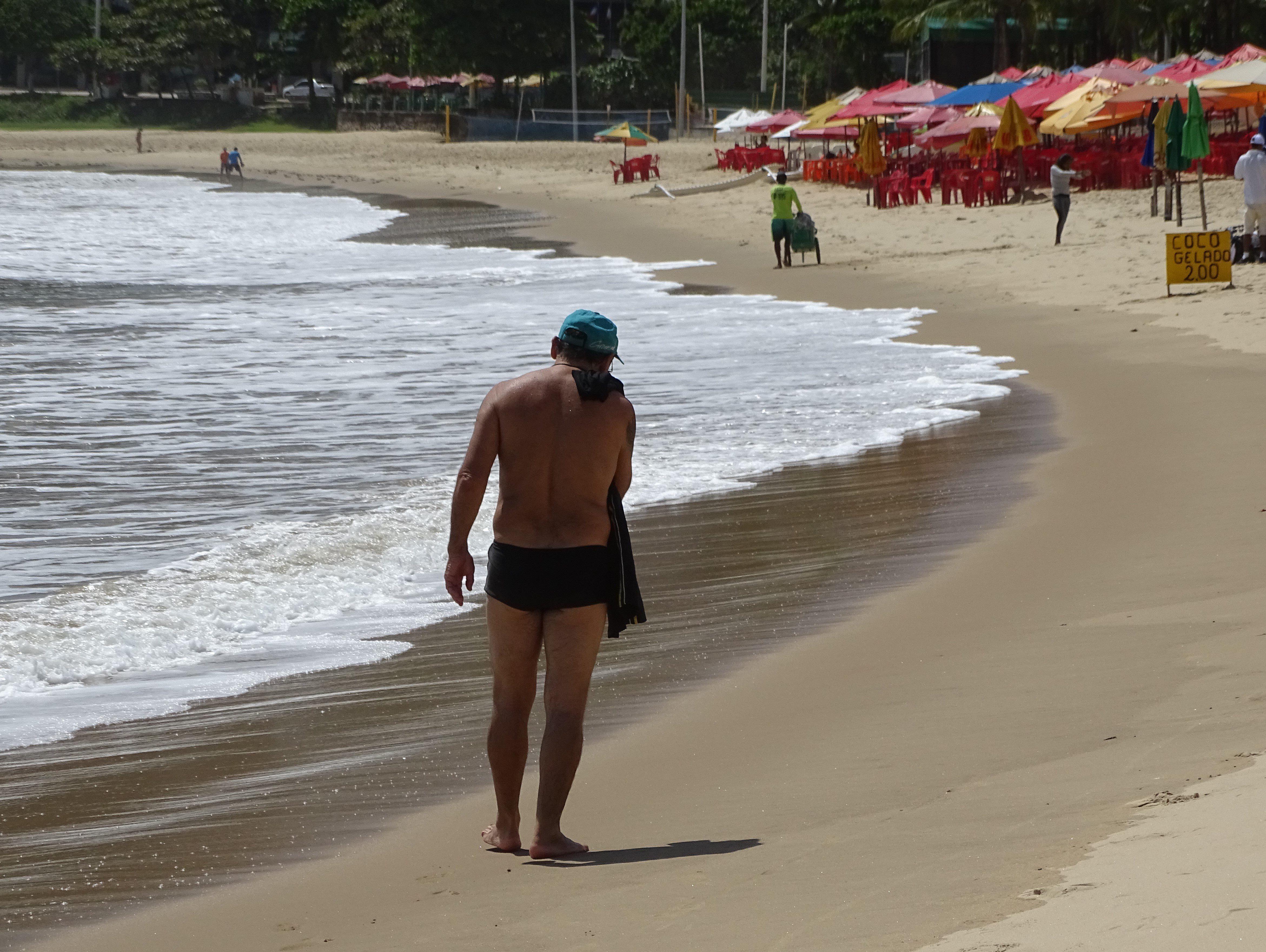 Fotografia de um homem caminhando na praia. À esquerda, as ondas fazem um semicírculo. Ao centro, o homem em destaque usa chapéu azul e tem a blusa pendurada no ombro. Ao fundo, na direita, algumas barracas de praia e um homem empurrando um carrinho de picolé
