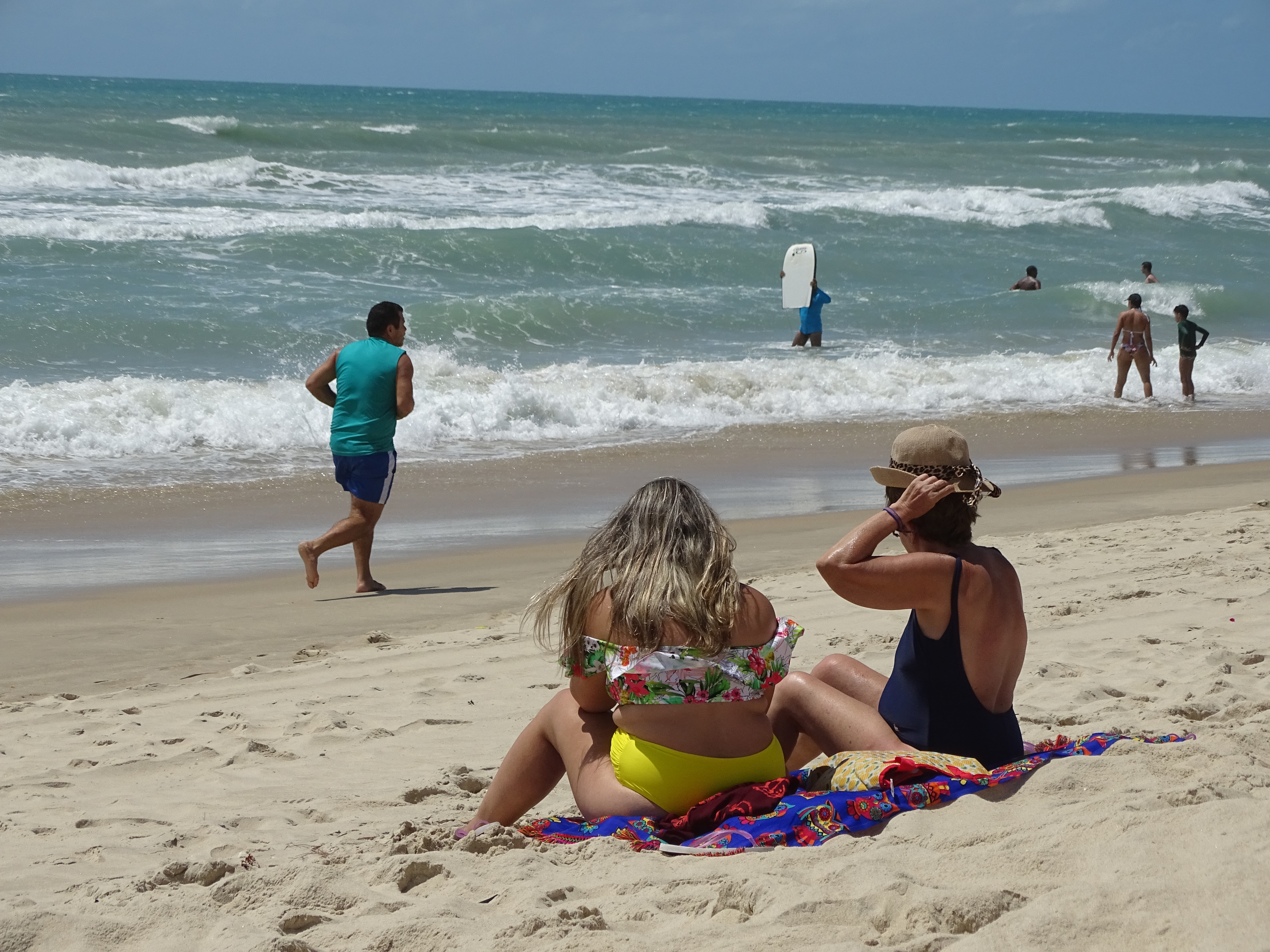 Fotografia da praia. Em primeiro plano, duas mulheres estão sentadas na areia olhando o mar. Uma delas coloca um chapéu na cabeça. Mais a frente um homem corre pela praia. Alguns banhistas estão dentro d'água