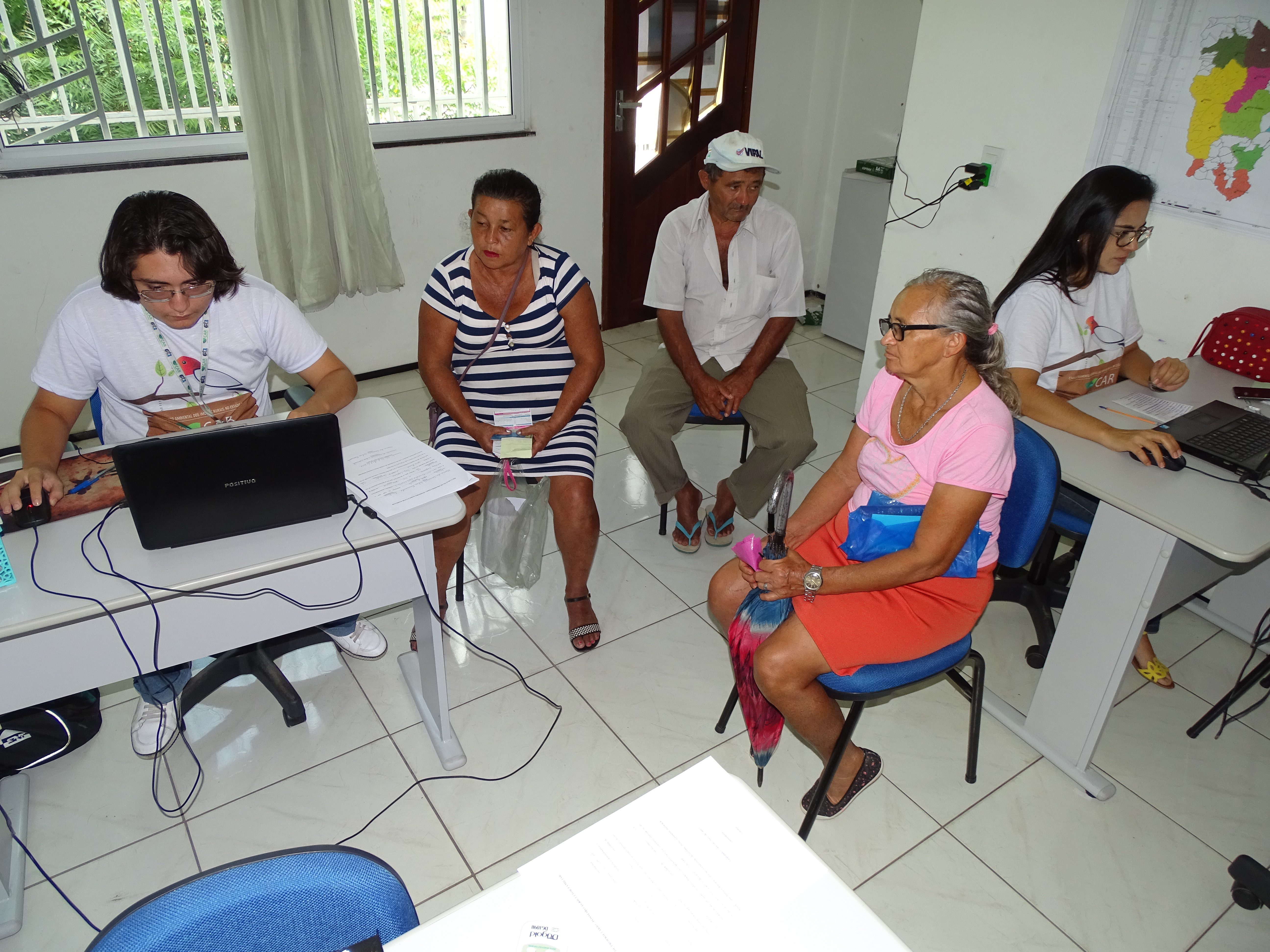 Fotografia da sala de um dos postos de atendimento do Cadastro Ambiental Rural (CAR). Na imagem à direita, um funcionário do CAR faz uma busca no notebook. Ao seu lado uma senhora de blusa listrada também olha para a tela do notebook. Um senhora segurando um guarda-chuva está sentada ao centro aguardando atendimento. No lado direito da imagem, uma funcionária do CAR também utiliza um notebook. Um senhor sentado ao fundo observa a funcionária enquanto aguarda atendimento.