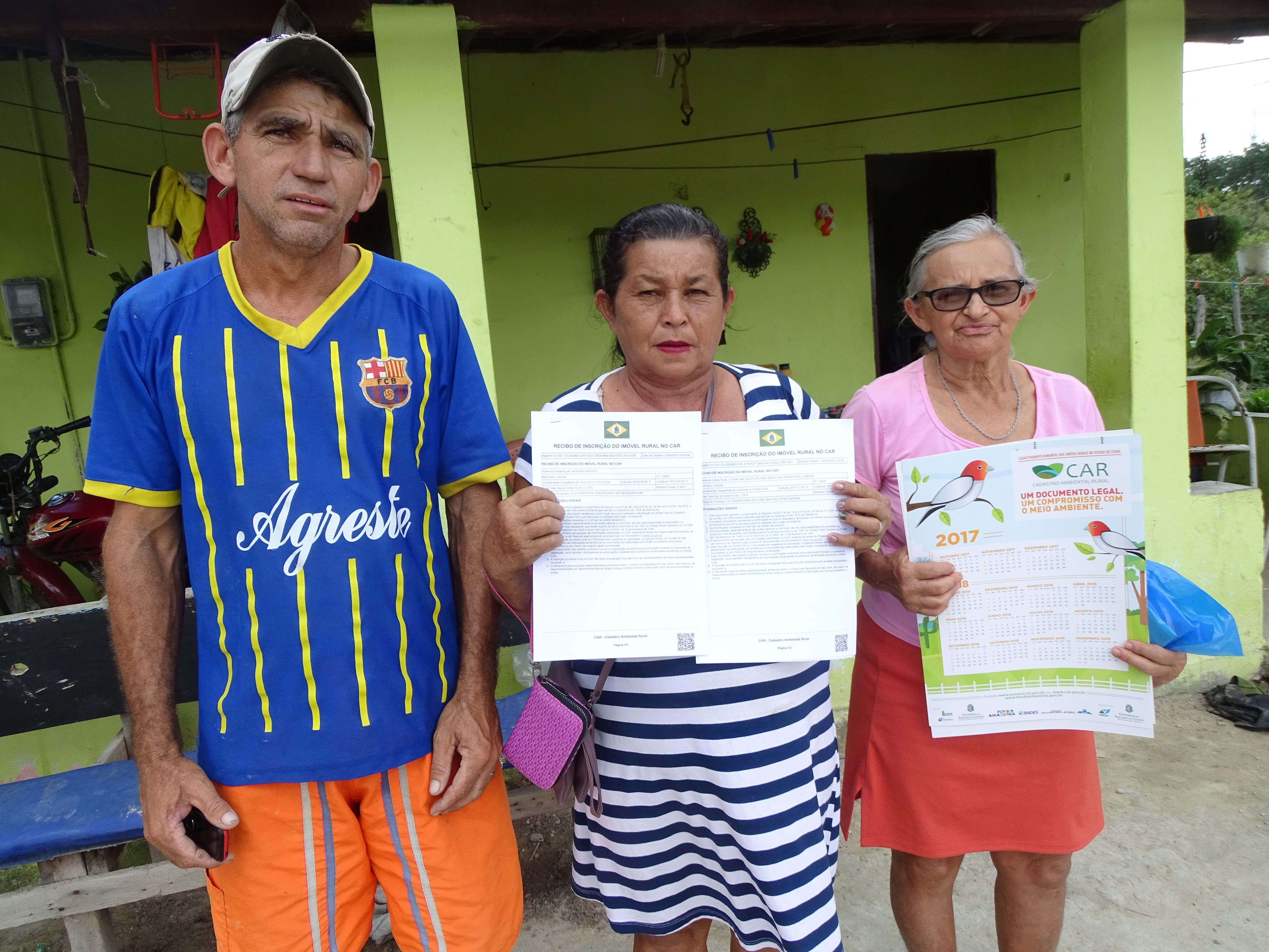 Fotografia. Da esquerda para a direita, um homem e duas mulheres. As mulheres expõem papéis do Cadastro Ambiental Rural. Os três estão em frente a uma casa de varanda com paredes verdes