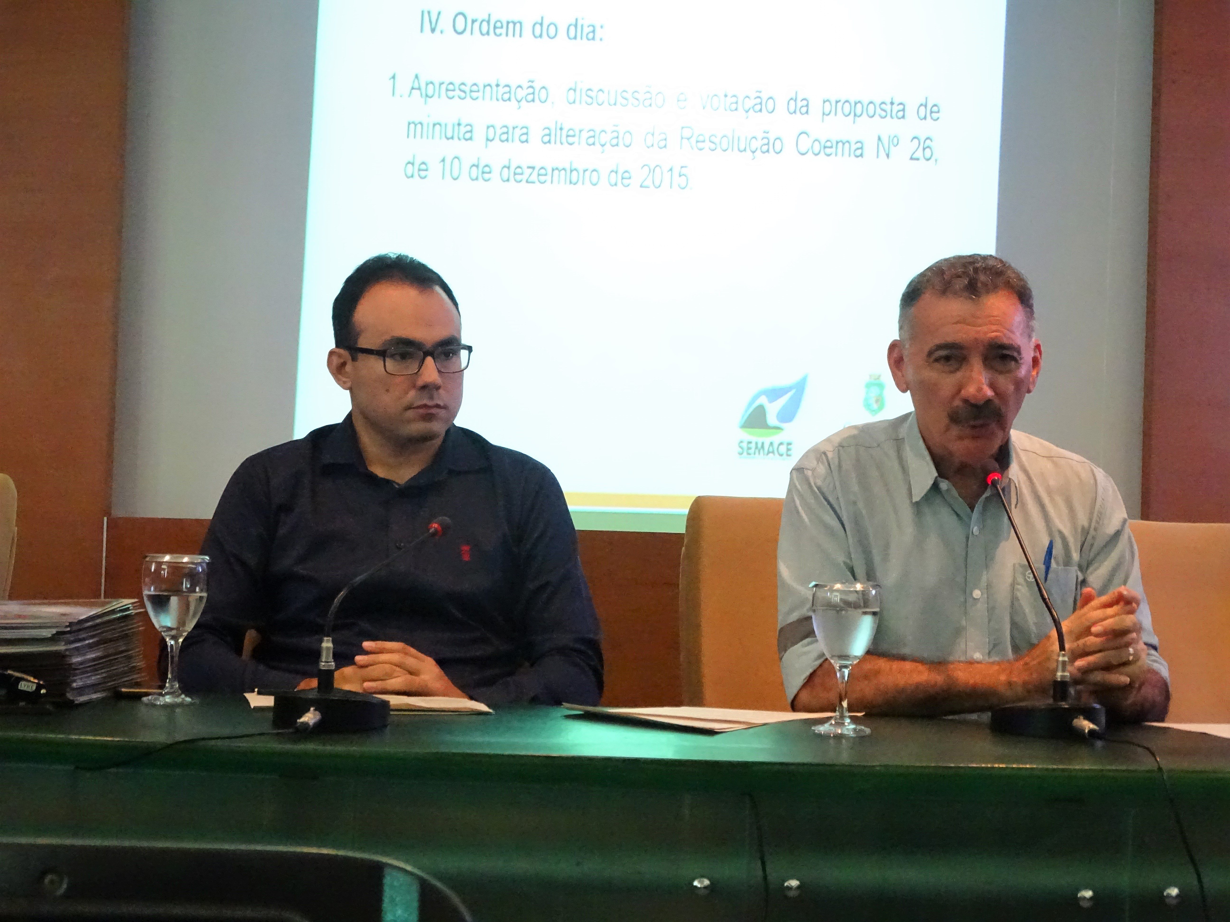 Fotografia da reunião do Coema. Na imagem, o secretário executivo do Coema e superintendente da Semace, Carlos Alberto Mendes, aparece à esquerda, ao lado do presidente do Coema, Arthur Bruno. Eles estão sentados na mesa principal do auditório atrás de seus respectivos microfones. Ao fundo uma apresentação em slides é apresentada. No slide está escrito: IV. Ordem do dia. 1. Apresentação, discussão e votação da proposta de minuta para a alteração da Resolução Coema Nº 26 de 10 de dezembro de 2015