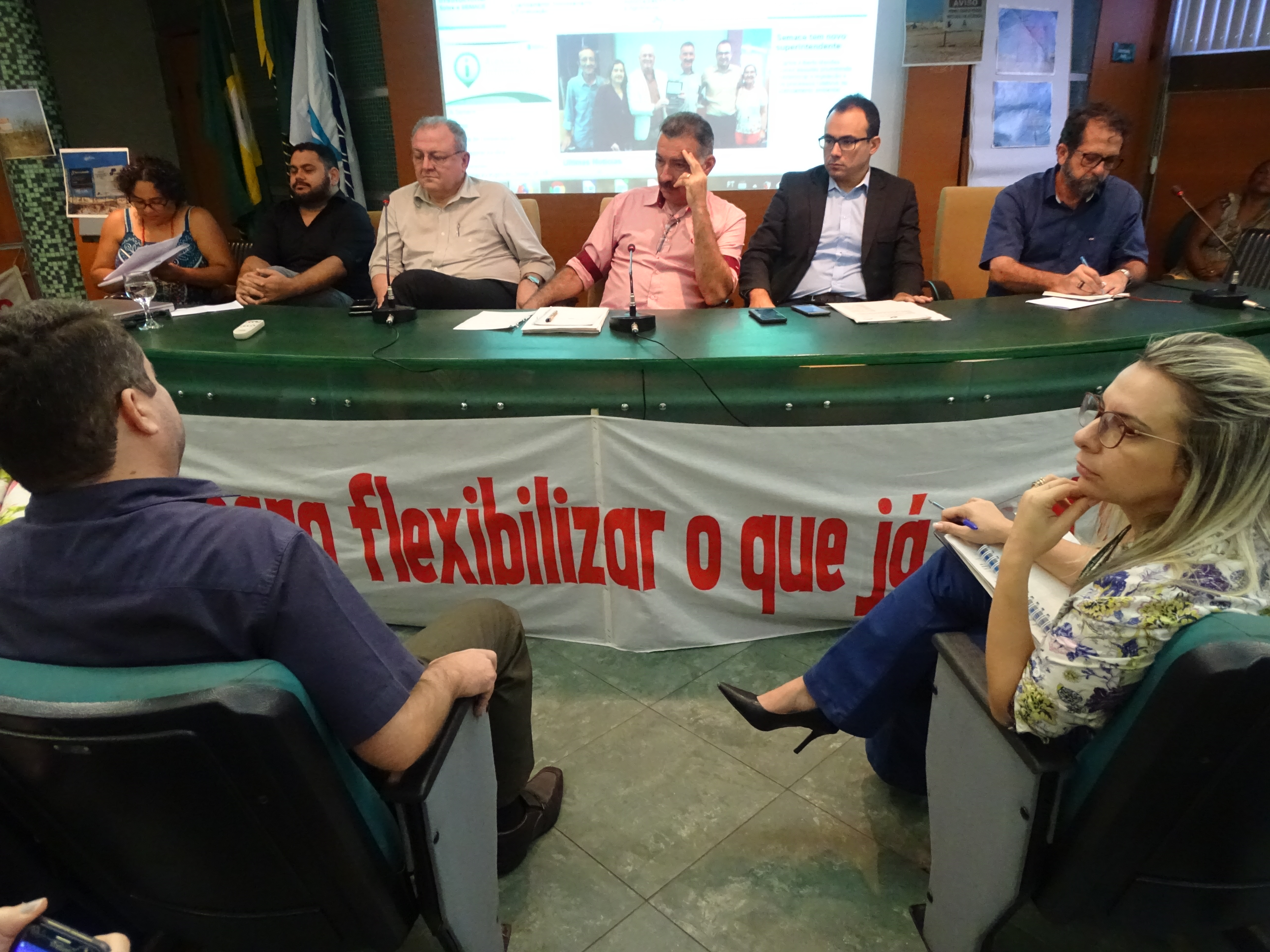 Fotografia da reunião do Coema em um auditório em tons de verde e marrom. Na bancada verde estão sentados seis representantes: cinco homens e uma mulher. Uma faixa com letras vermelhas está colada na frente da bancada. Na parte visível está escrito: flexibilizar o que já. Uma imagem projetada ao fundo mostra a tela inicial do site da Semace