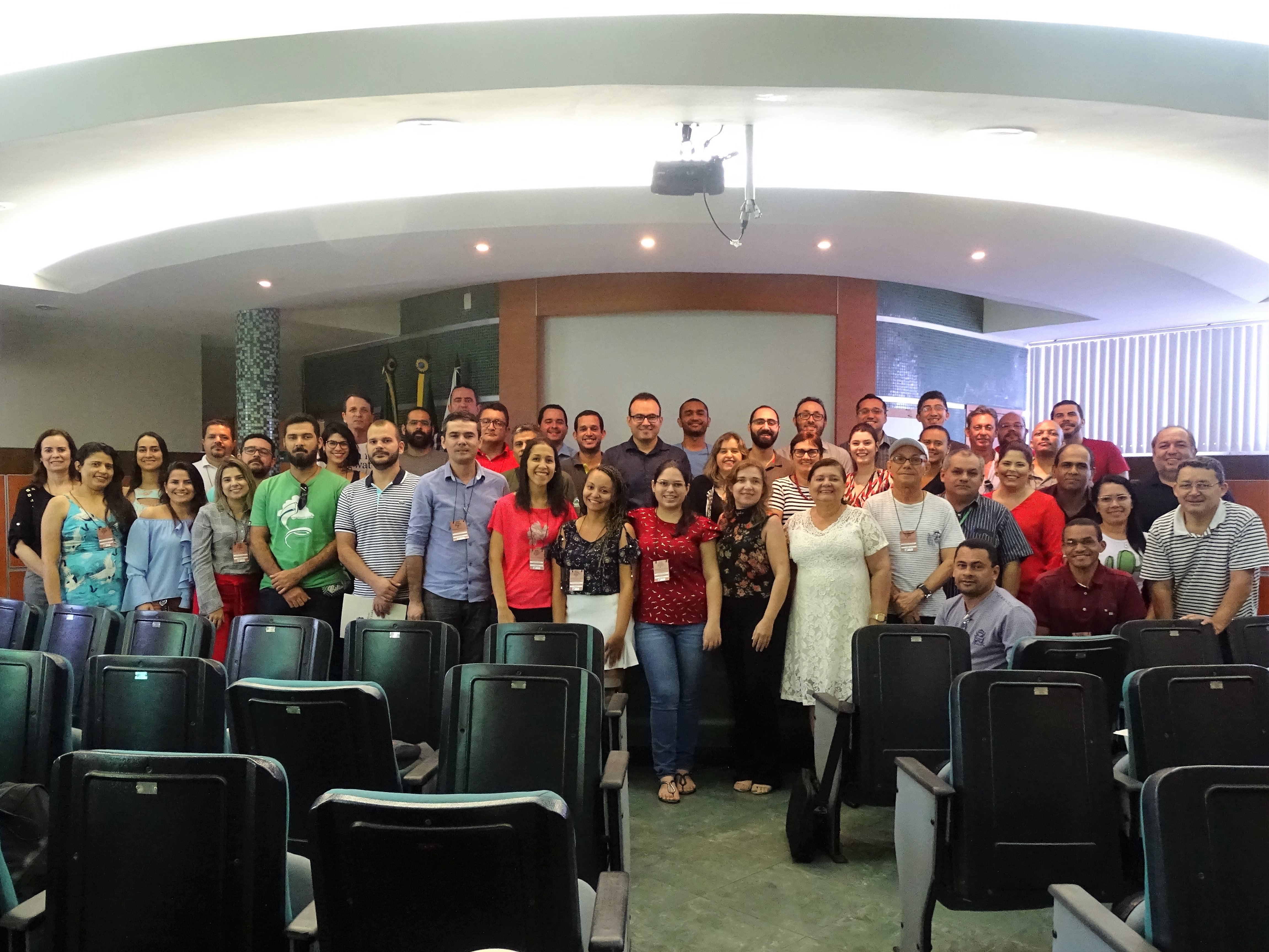 Fotografia em grupo dos participantes do Curso Interação. A foto foi tirada no auditório da Semace. À frente estão os assentos, em tons de verde. No centro o grupo de pessoas. Atrás, a parede verde com um espaço em branco onde fica a imagem do projetor