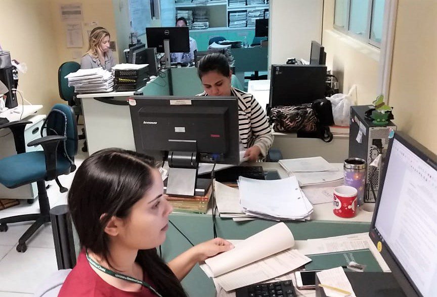 Fotografia de uma sala em tons de verde, onde estão dispostas sete mesas e cadeiras, também em verde. Sob as mesas, estão computadores e uma grande quantidade de documentos. Quatro mulheres estão sentadas em frente aos computadores, analisando as telas ou papéis