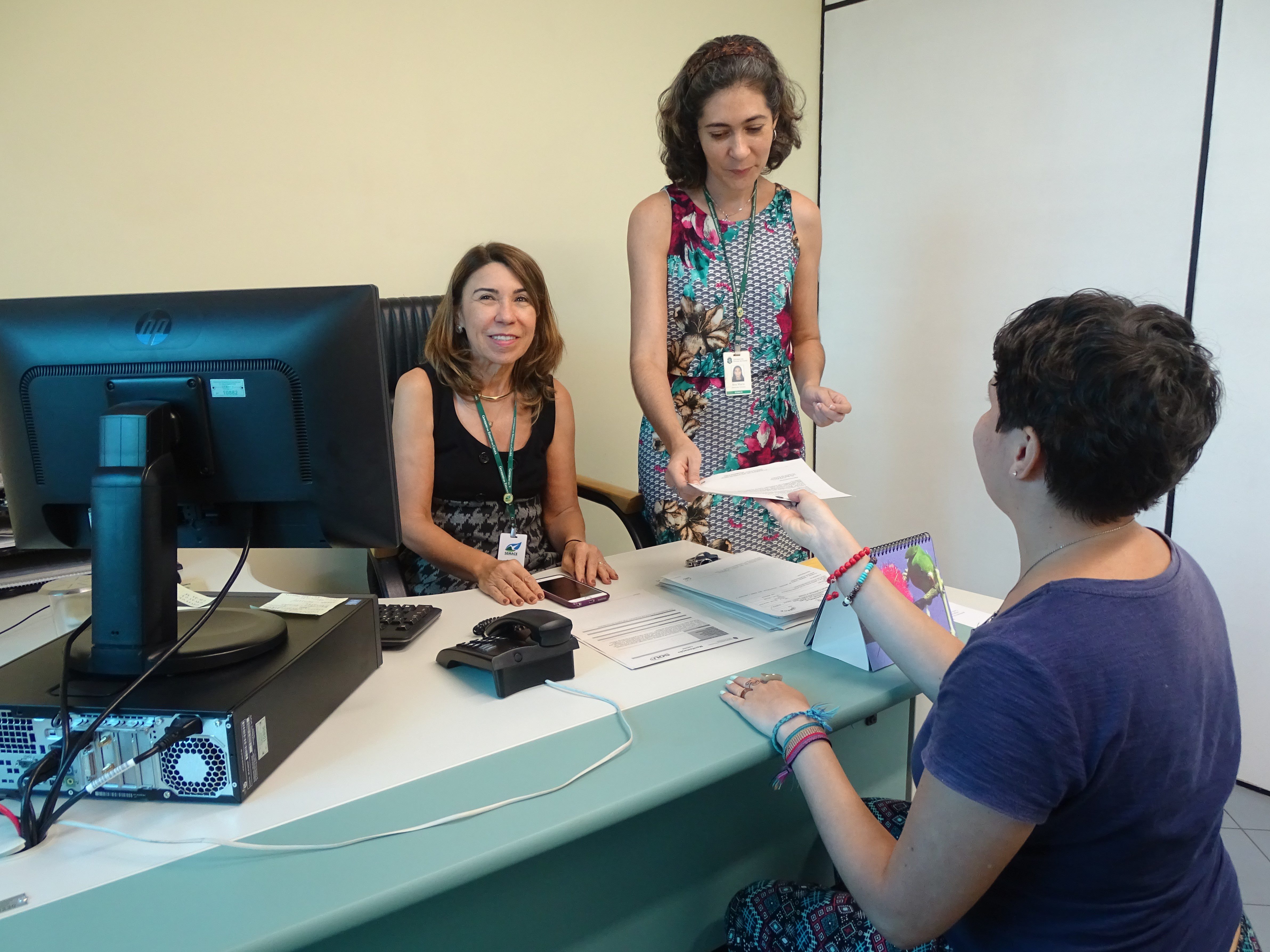 Na imagem, três mulheres estão na sala da ouvidoria da Semace. As paredes têm tons de verde. Duas estão atrás de uma mesa onde fica um computador. Uma delas entrega um papel à que está do outro lado da mesa