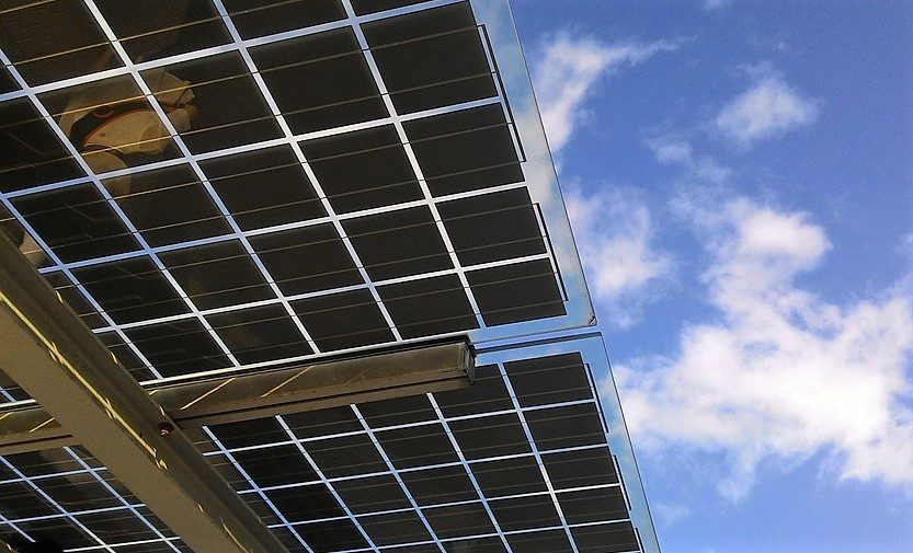 Fotografia de um painel de energia solar visto de baixo. Ao fundo, um céu azul com algumas nuvens brancas