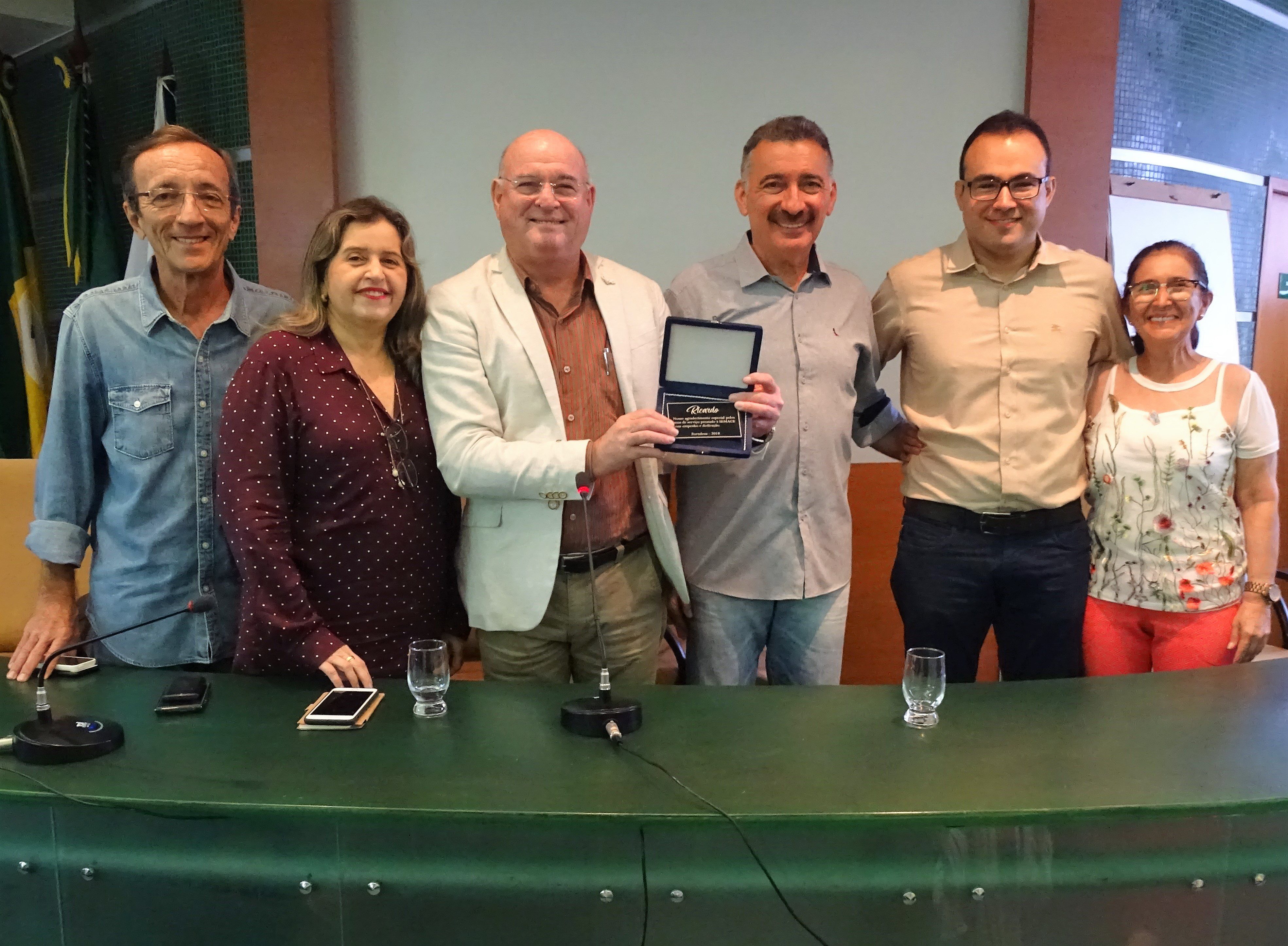Fotografia do momento da posse. Na imagem estão seis pessoas, quatro homens e duas mulheres, atrás da bancada verde do auditório da Semace. Ao centro, da esquerda para a direita, estão o até então superintendente da Semace, Ricardo Araújo; o secretário da Sema, Arthur Bruno e o novo superintendente, Carlos Alberto Mendes.