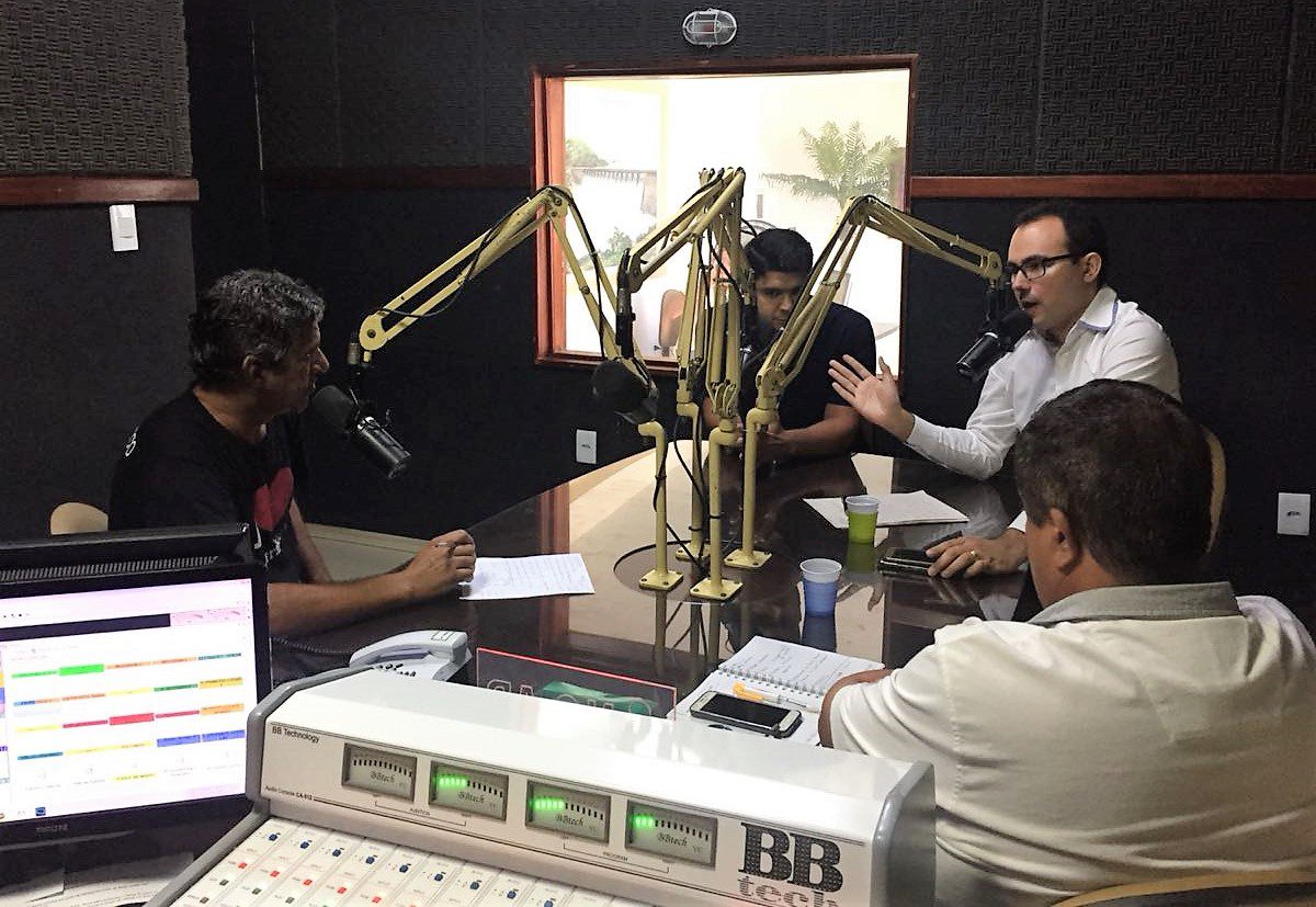 Fotografia da sala da rádio em Quixeramobim, com paredes escuras. Em primeiro plano estão os equipamentos de controle da rádio. Ao centro, o superintendente da Semace, Carlos Alberto Mendes está sentado em uma mesa com três outros homens, enquanto fala ao microfone. Dois dos outros presentes também estão em frente à microfones. Ao fundo, uma janela de vidro mostra algumas plantas