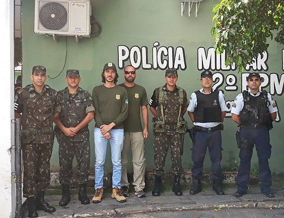 Fotografia. Sete homens, usando roupas do exército, polícia e fiscalização. Ao fundo, em uma parede verde, está escrito: Polícia Militar. 2º BPM