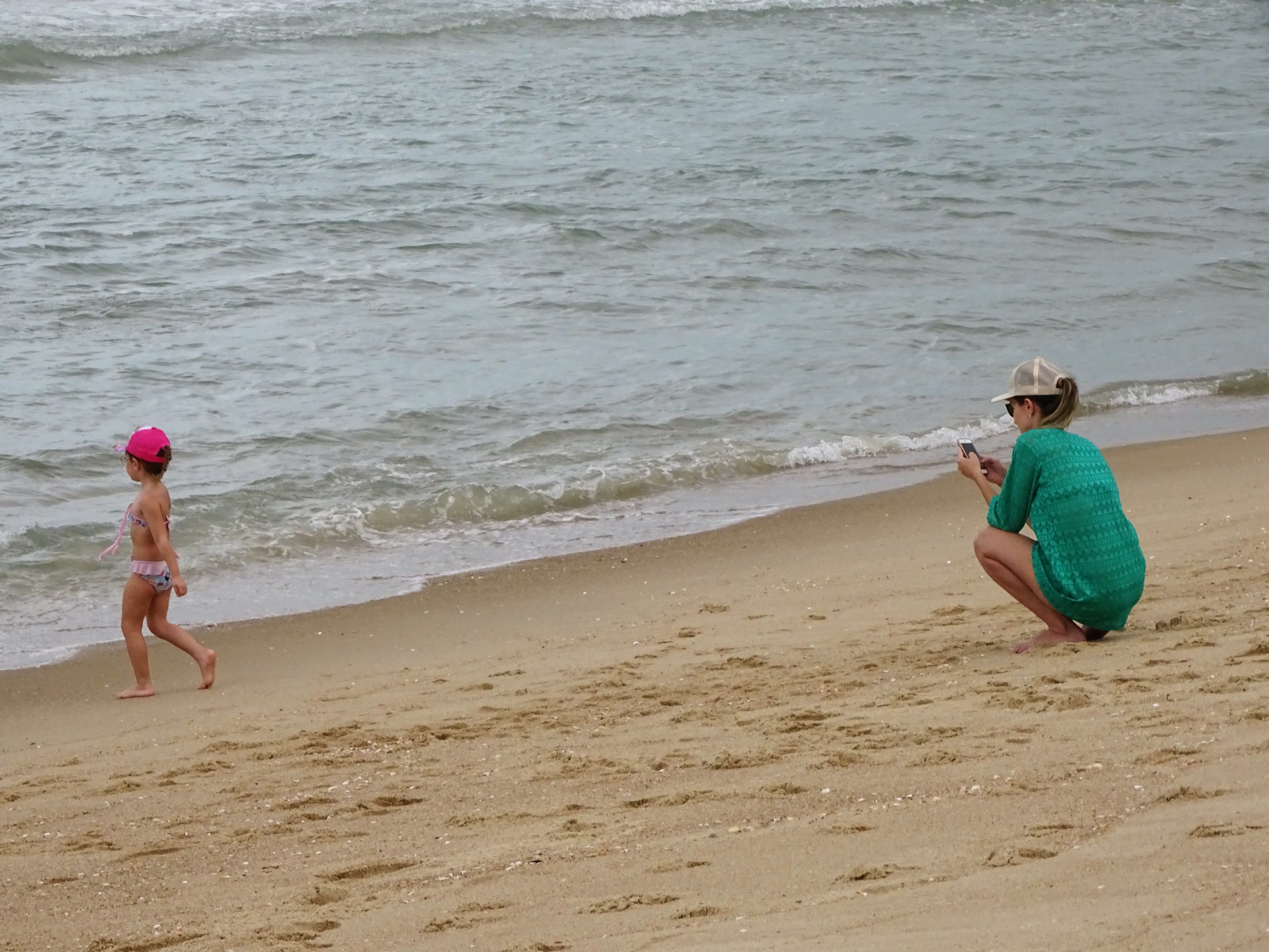 Fotografia de mulher observando uma criança brincando na praia, próxima ao mar