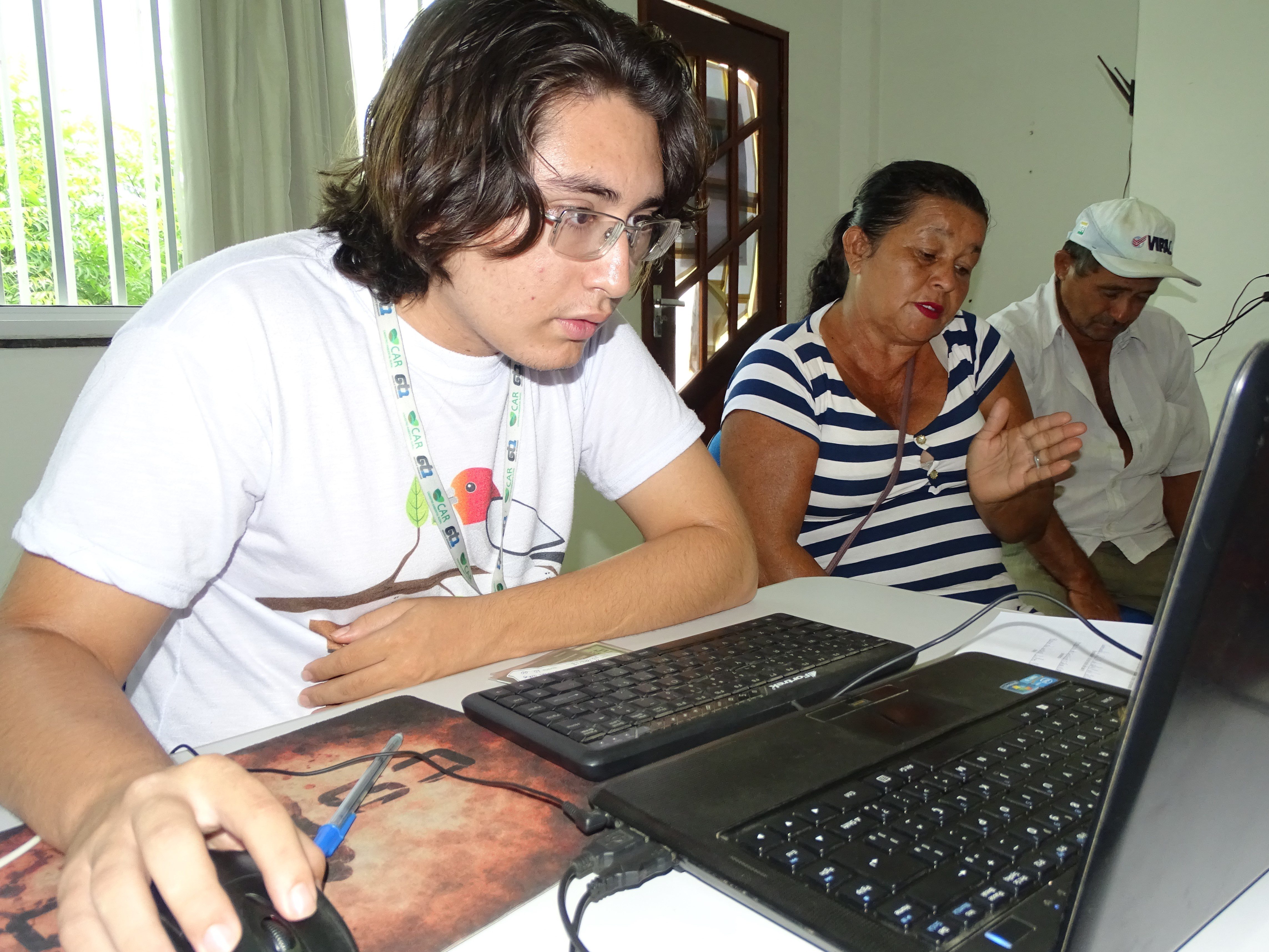 Fotografia do atendimento do CAR sendo executado. Na imagem, tirada na digonal,um funcionário do CAR aparece em primeiro plano. Ele segura um mouse enquanto olha para a tela do notebook à sua frente. Ao seu lado, em segundo plano, uma senhora de blusa listrada fala e gesticula. Ao fundo um senhor está sentado