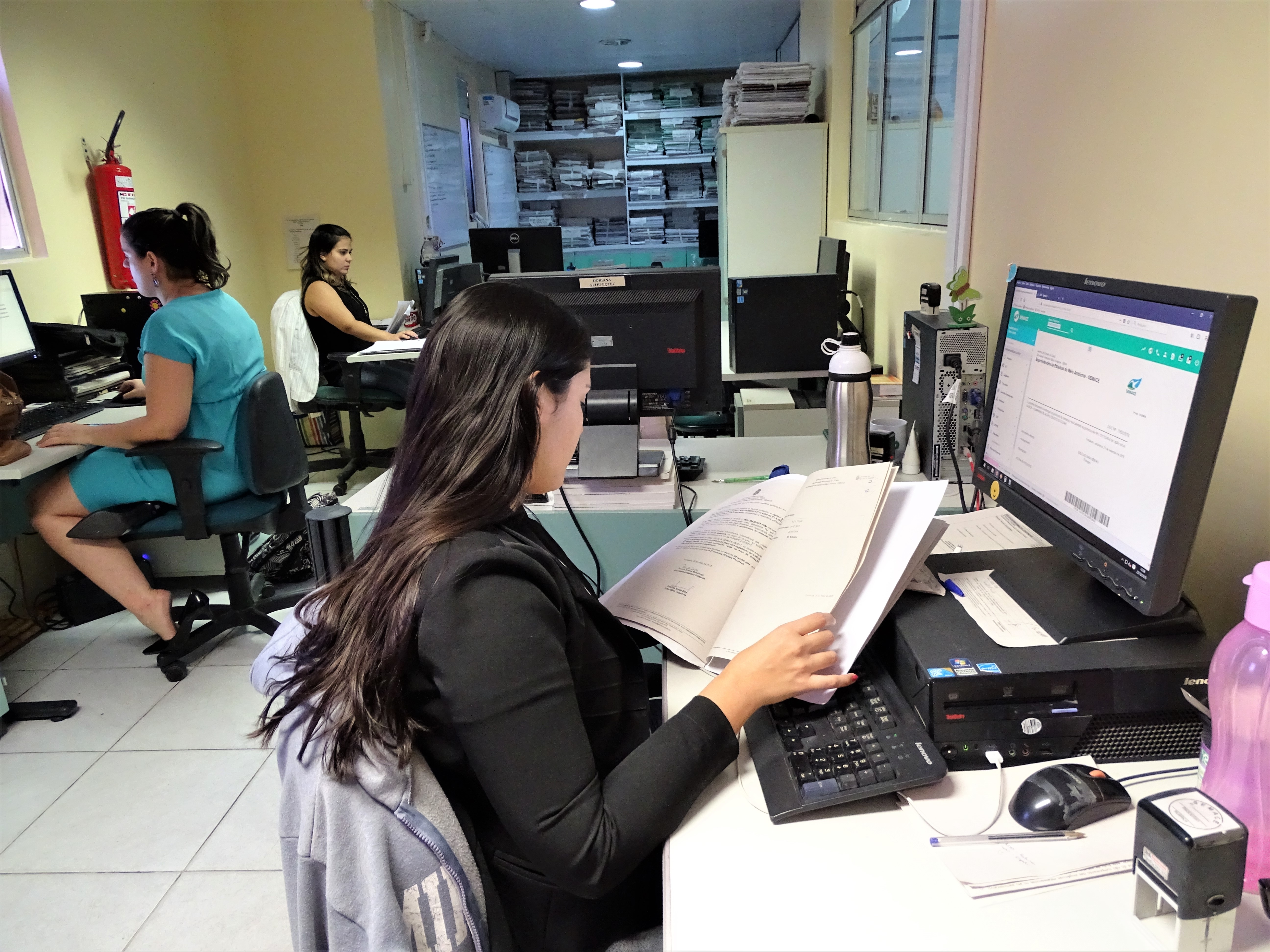 fotografia da sala da geiju. Em primeiro plano, uma mulher sentada a frente de um computador verifica alguns papéis